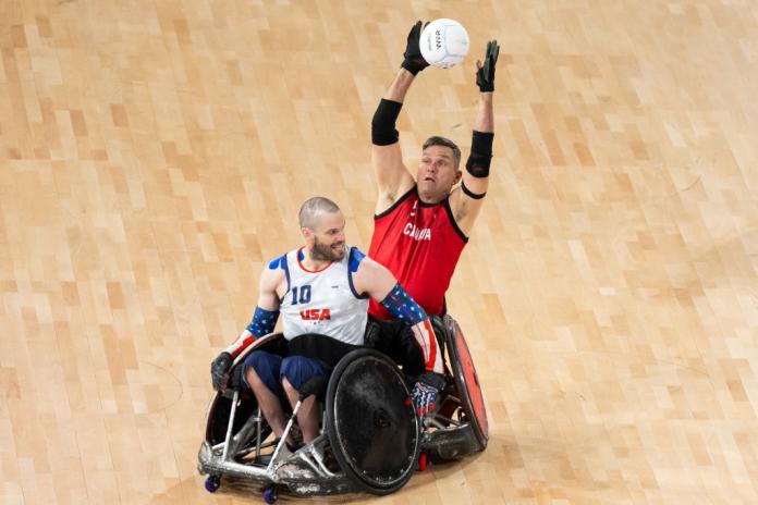 A male wheelchair rugby player is reaching up to grab a ball 